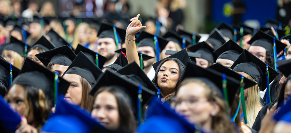 Graduates in black caps and gowns sit in rows, with one hand raised making a celebratory gesture.