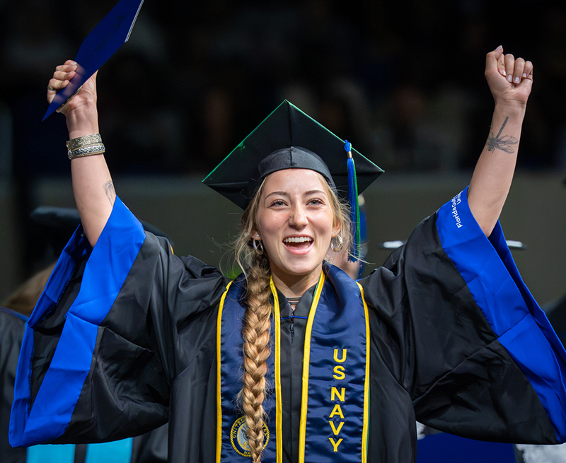 Graduate in cap and gown with US Navy stole raises both arms, holding a diploma folder in one hand.
