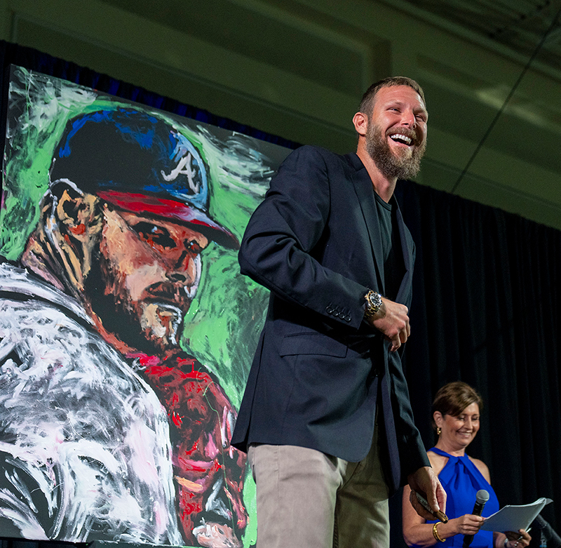 Person stands near a colorful painting of a baseball player during an auction event.