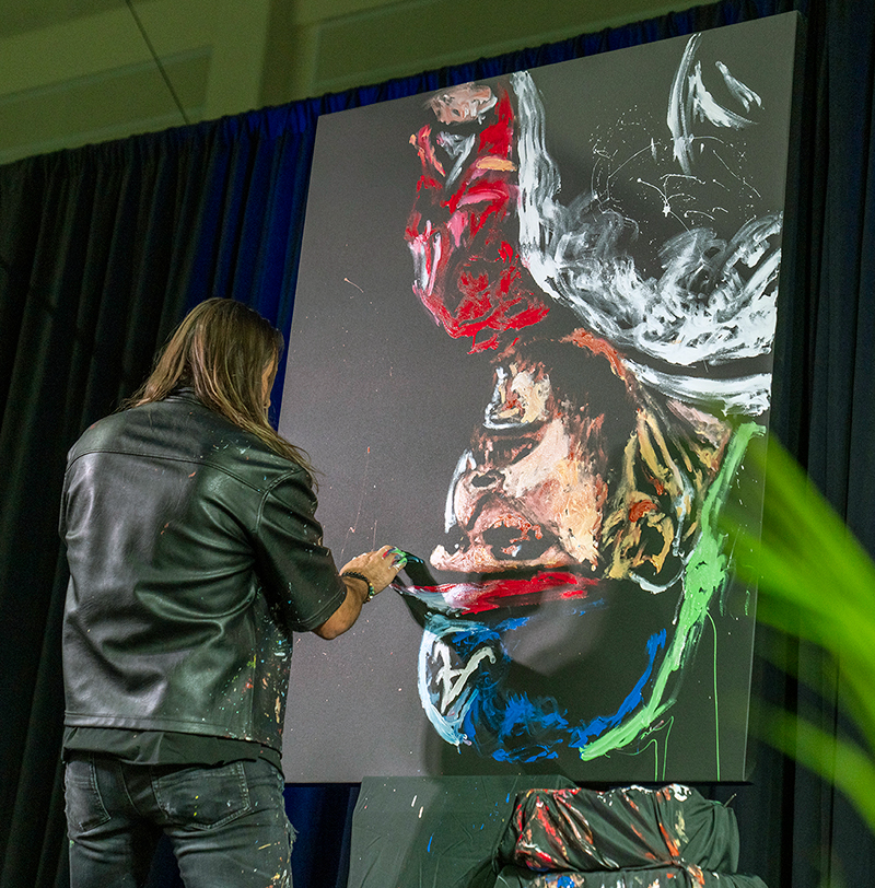 Artist paints a colorful abstract portrait on a large black canvas during a live event.