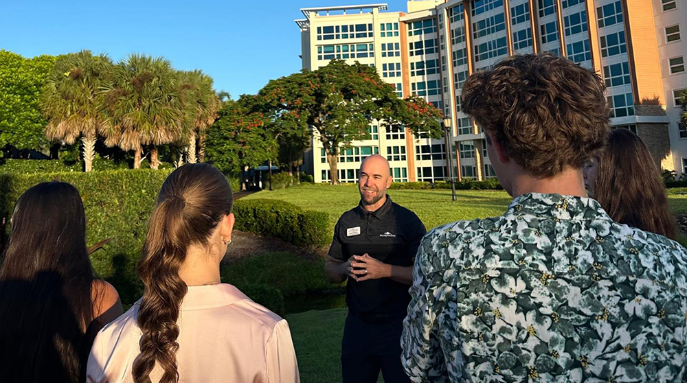 Person speaking outdoors to a small group near a landscaped lawn with a large building in the background.