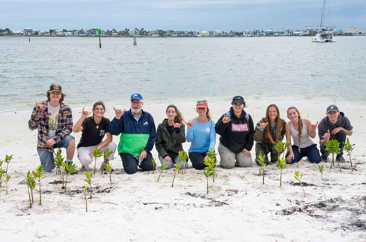 Nine people kneel on the beach in front of small plants, making the FGCU Wings Up gesture