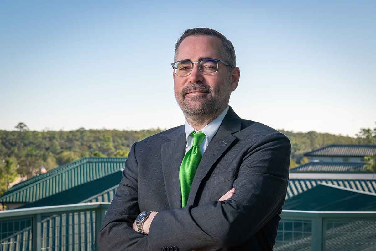 A man in a dark grey suit, glasses and a green tie, stands outdoors with folded arms and a railing and green, metal rooftops visible behind him