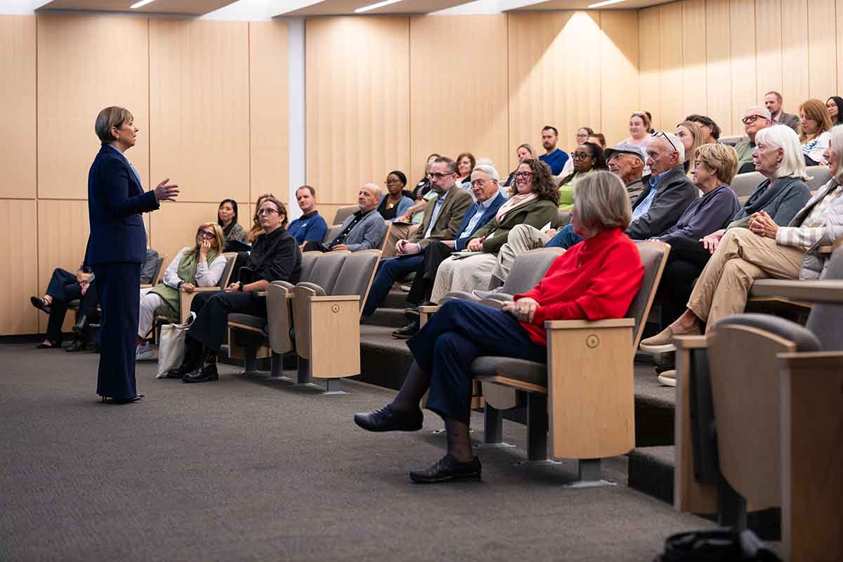 A woman in a navy blazer and pale blue blouse stands, speaking to a tiered classroom of people