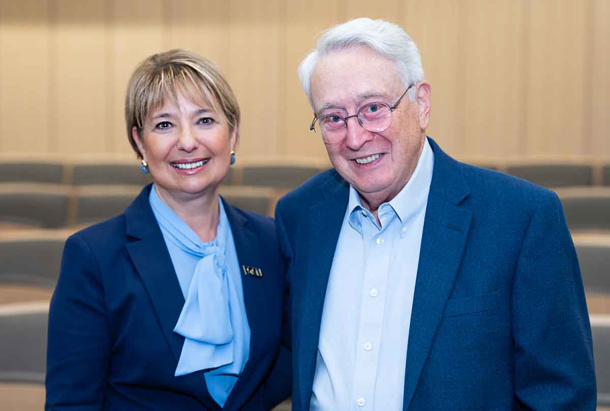 A woman with short, blonde hair and a navy blazer over a pale blue blouse stands smiling next to a man in a blue blazer and light blue button-down shirt