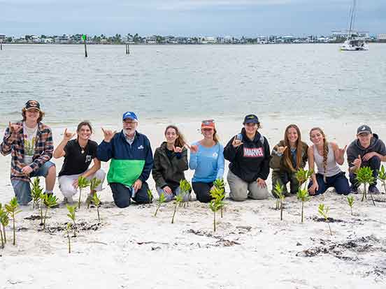 Nine people kneel on the beach in front of small plants, making the FGCU Wings Up gesture