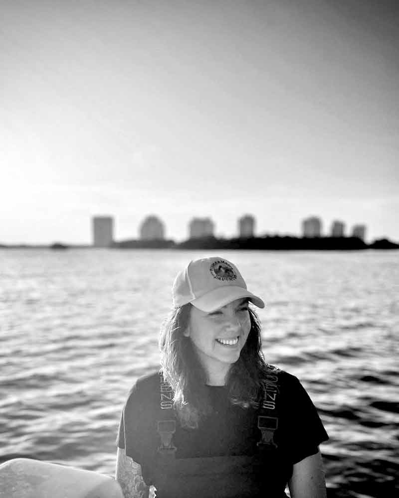 A black and white photo of a woman wearing a white cap with a round, Fisherman's' Daughter logo and overalls over a dark shirt. The background shows the water and a skyline behind her, as she appears to be on a boat