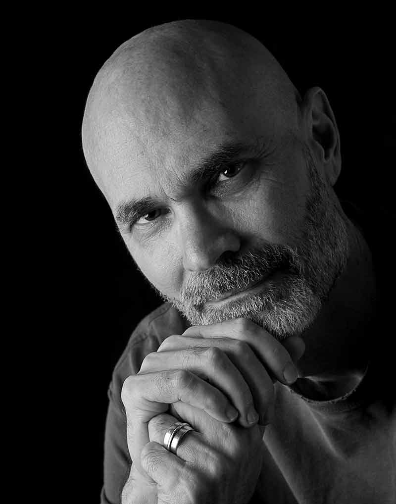 A black and white portrait of a bald man with grey and white moustache and beard and his hands clasped in front of him, wearing a ring on his pinky