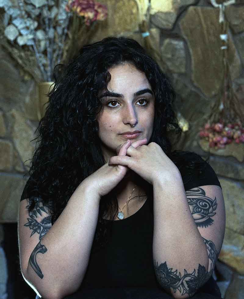 A color portrait of a woman with long, black air, tattoos on both arms, clasping her hands under her chin and looking into the distance