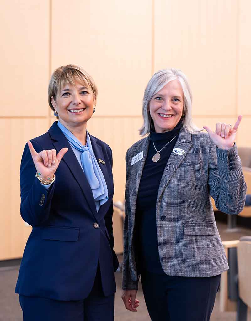 Two women stand, dressed in business attire, smiling and making the FGCU Wings Up gesture