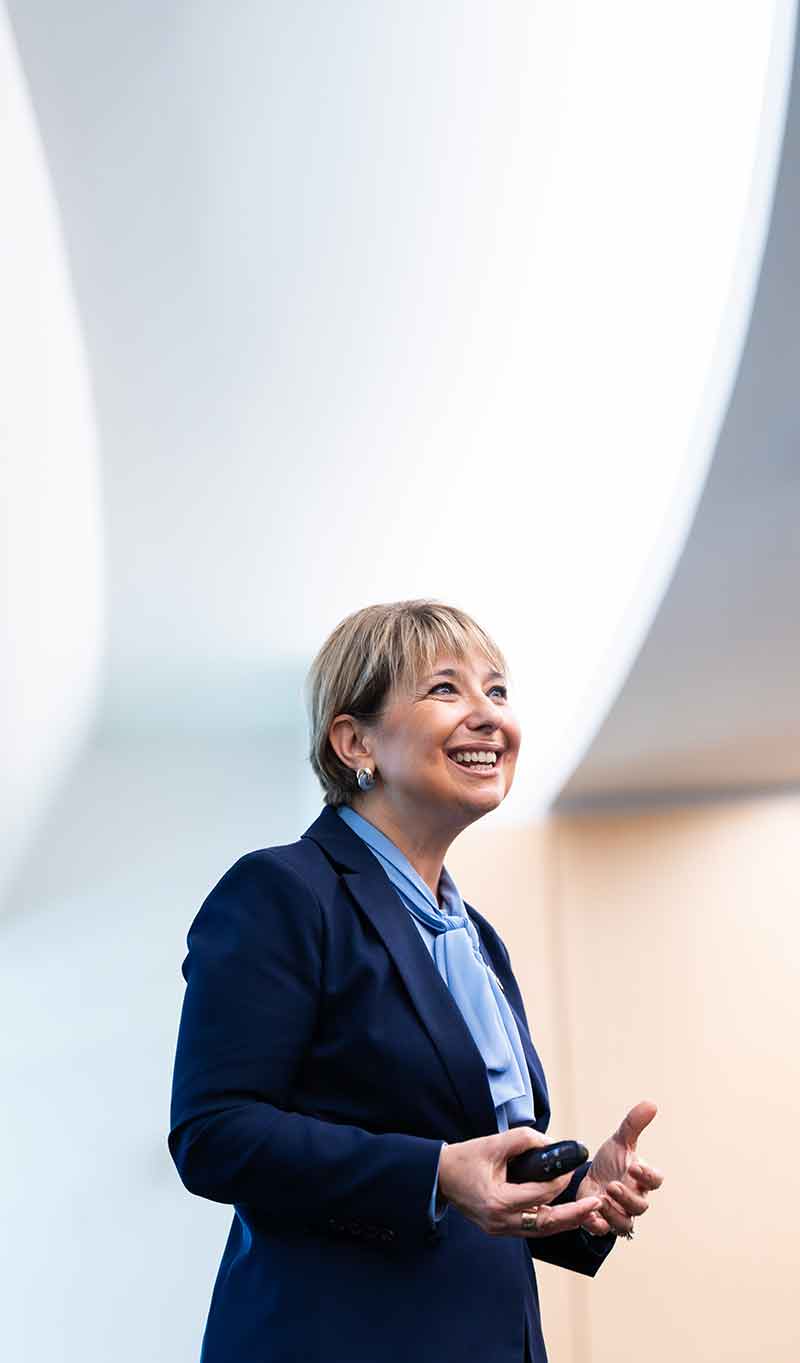 A woman with short, blonde hair and a navy blazer over a pale blue blouse stands smiling in a room with a white ceiling in a dramatic arch over her head