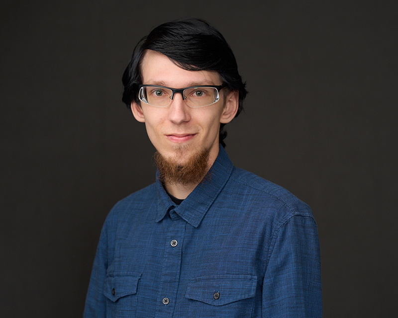Person wearing a blue button-up shirt posed against a dark studio background.