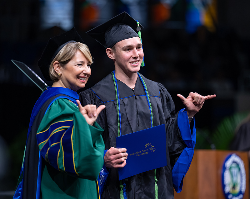 Graduate in cap and gown holding diploma next to faculty member in academic regalia at ceremony.