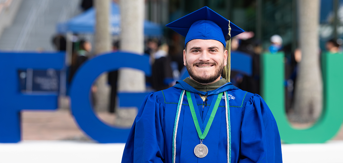 Graduate in blue cap and gown with green honor cords and medal standing in front of large FGCU letters.