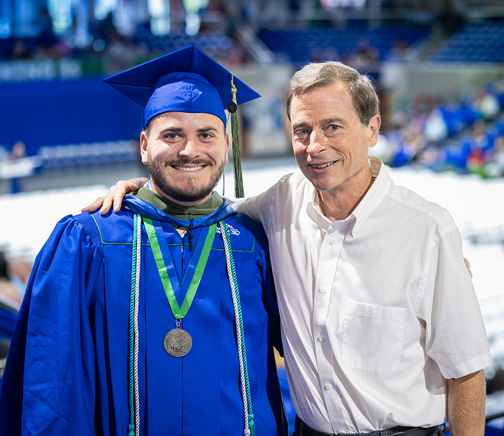 Graduate in blue cap and gown with green honor cords and medal standing beside a person in white shirt.