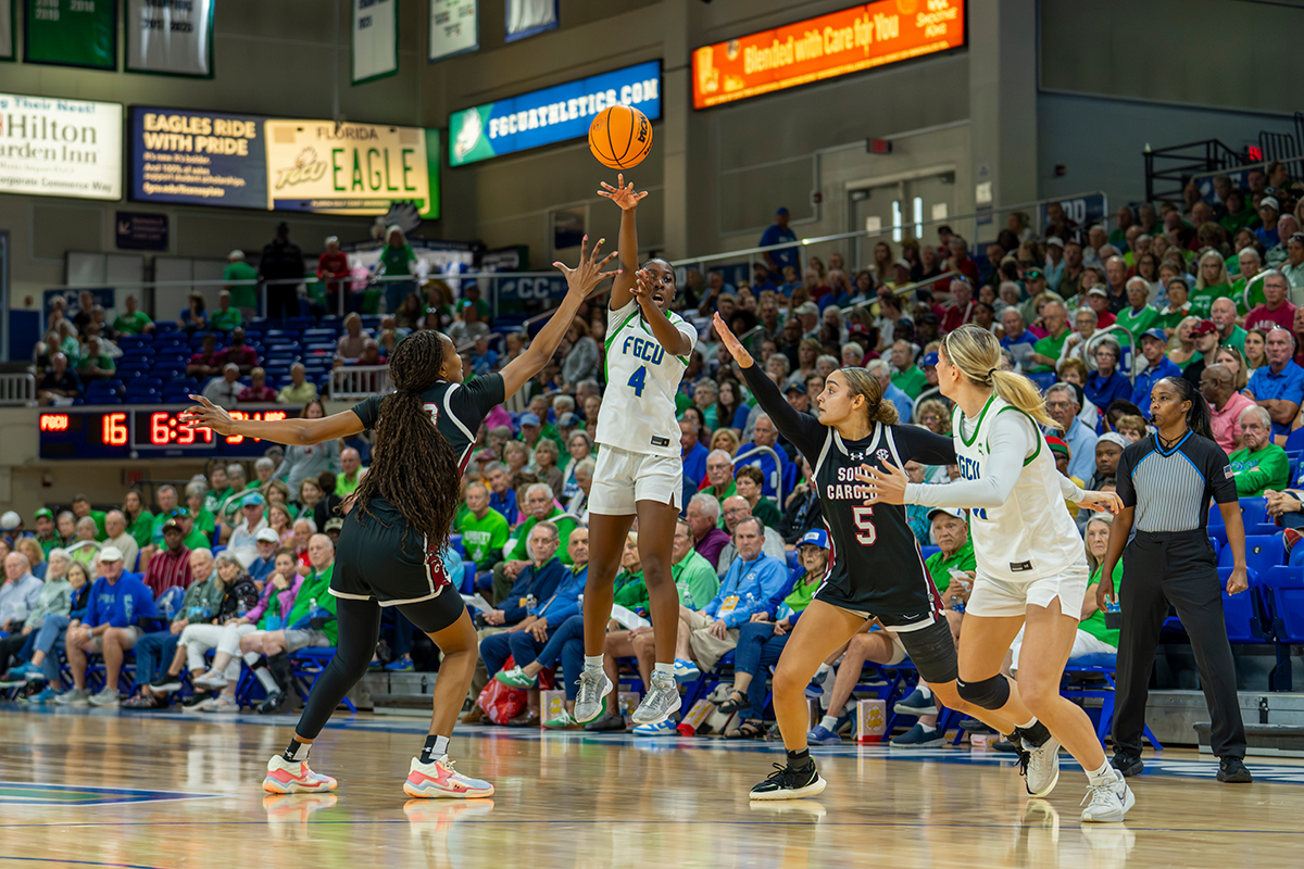 FGCU basketball player shoots while defended by South Carolina players during a game in a crowded arena.