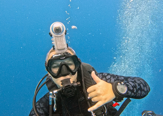 Scuba diver underwater with a mounted camera giving a Wings Up hand gesture amid rising bubbles.
