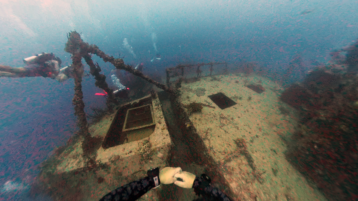 Diver explores a submerged shipwreck with other divers visible in the background underwater.