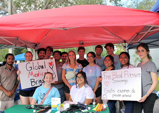 Group under red canopy holding signs for Global Medical Brigades and free blood pressure screenings in English and Spanish.