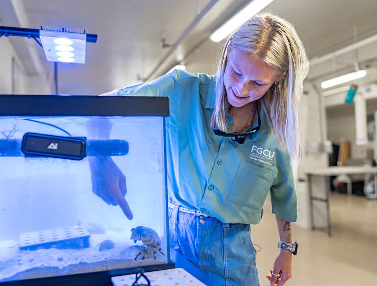 A young, blonde woman in a light green button-up shirt has her right arm submerged in a fish tank to her elbow and is pointing at a crab in the tank