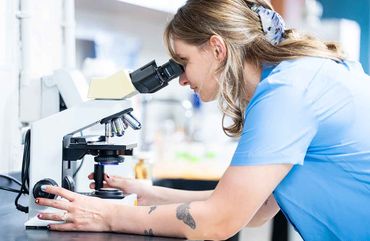 A young woman in a blue, short-sleeved shirt looks into a microscope