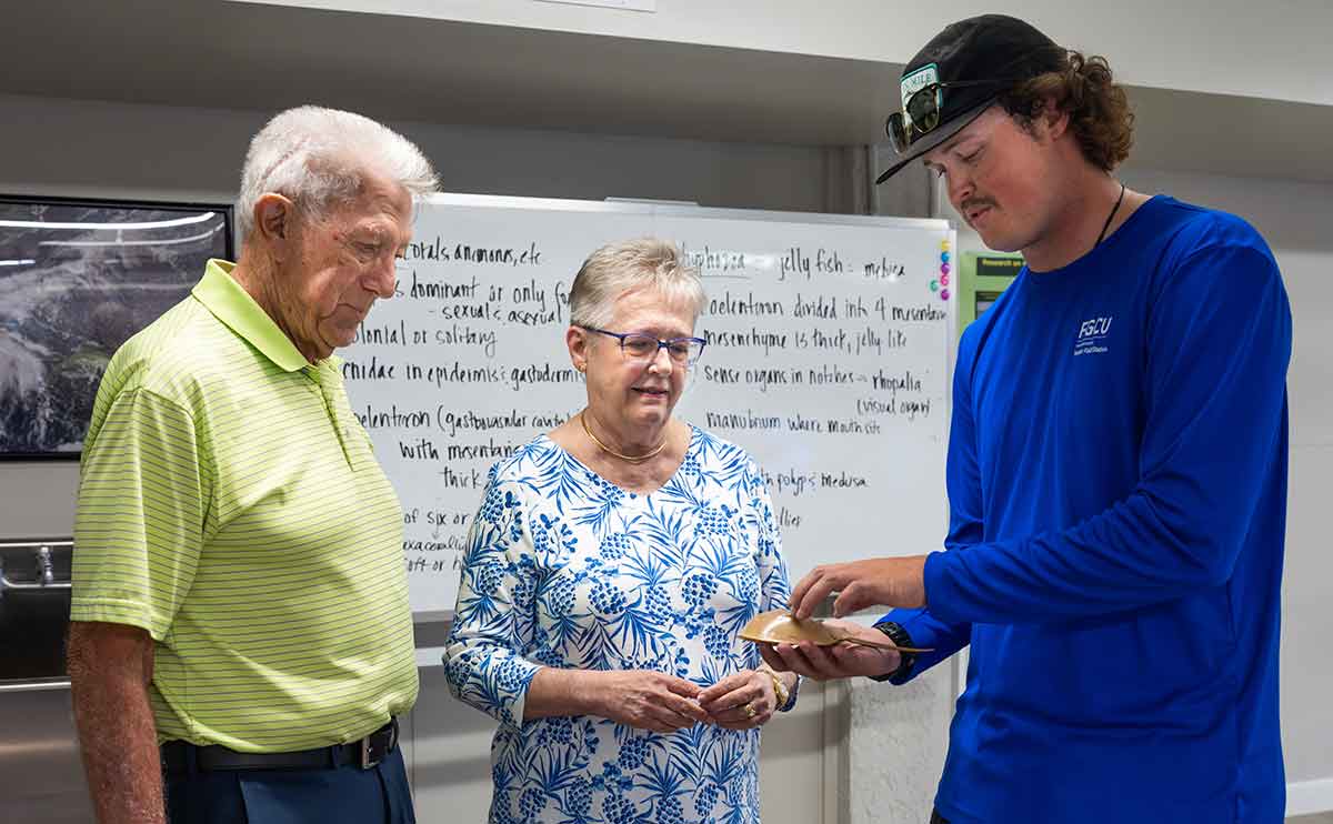 An older man in a lime green polo and an older woman in a patterned blue-and-white shirt look at an object in the hand of a tall, young man in a long-sleeved, bright blue shirt and ball cap