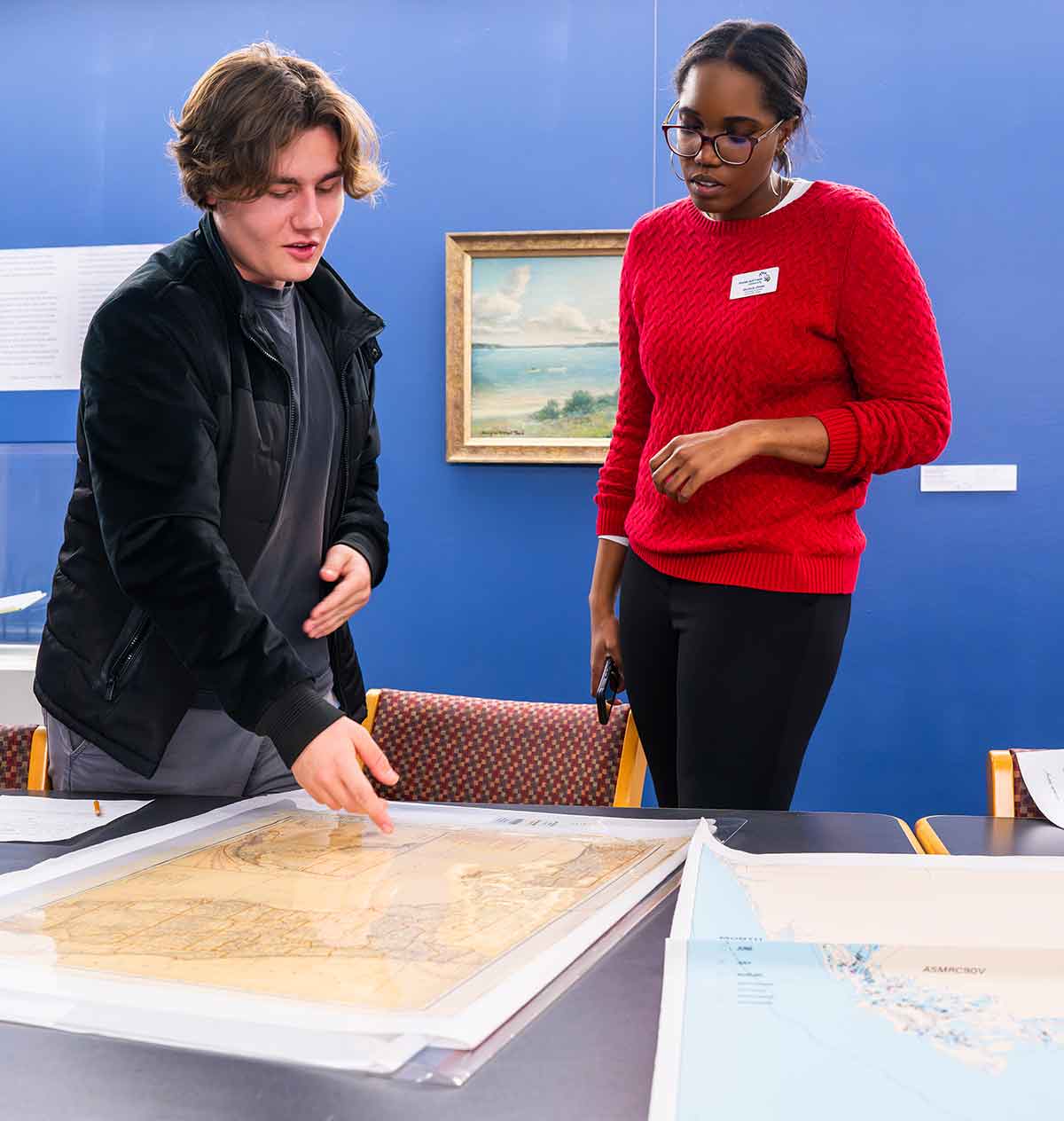 A young man in a black jacket and a woman in a red sweater stand, looking at an old map on a table