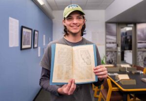 A young man in a light green FGCU hat holds a slim volume with print on it in front of his chest