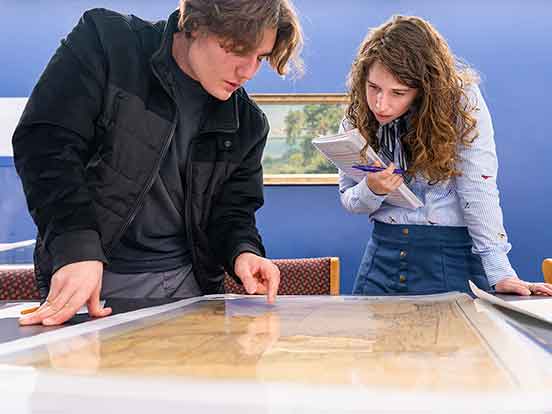 A young man in a black jacket and a woman in a blue button-down shirt and skirt stand, looking at an old map on a table