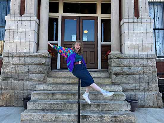 A young woman dressed in 1980s style clothing appears to slide down a railing in front of an old building with stone steps