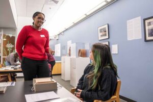 A woman in a red sweater stands, talking to a seated young woman with long, dark brown hair with green tips