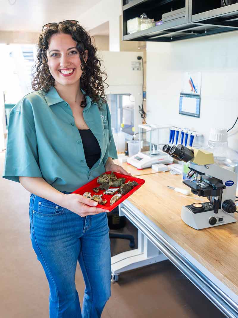 A woman in a green shirt over a black tank top and jeans holds a red tray of oysters in a laboratory