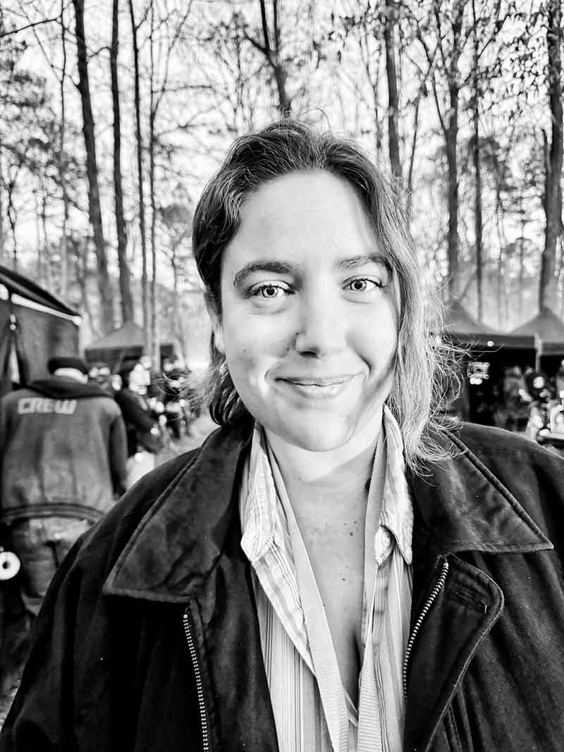 A black and white photo of a woman, her hair pulled back, wearing a black jacket over a white shirt, with people in the background and one wearing a shirt that reads Crew
