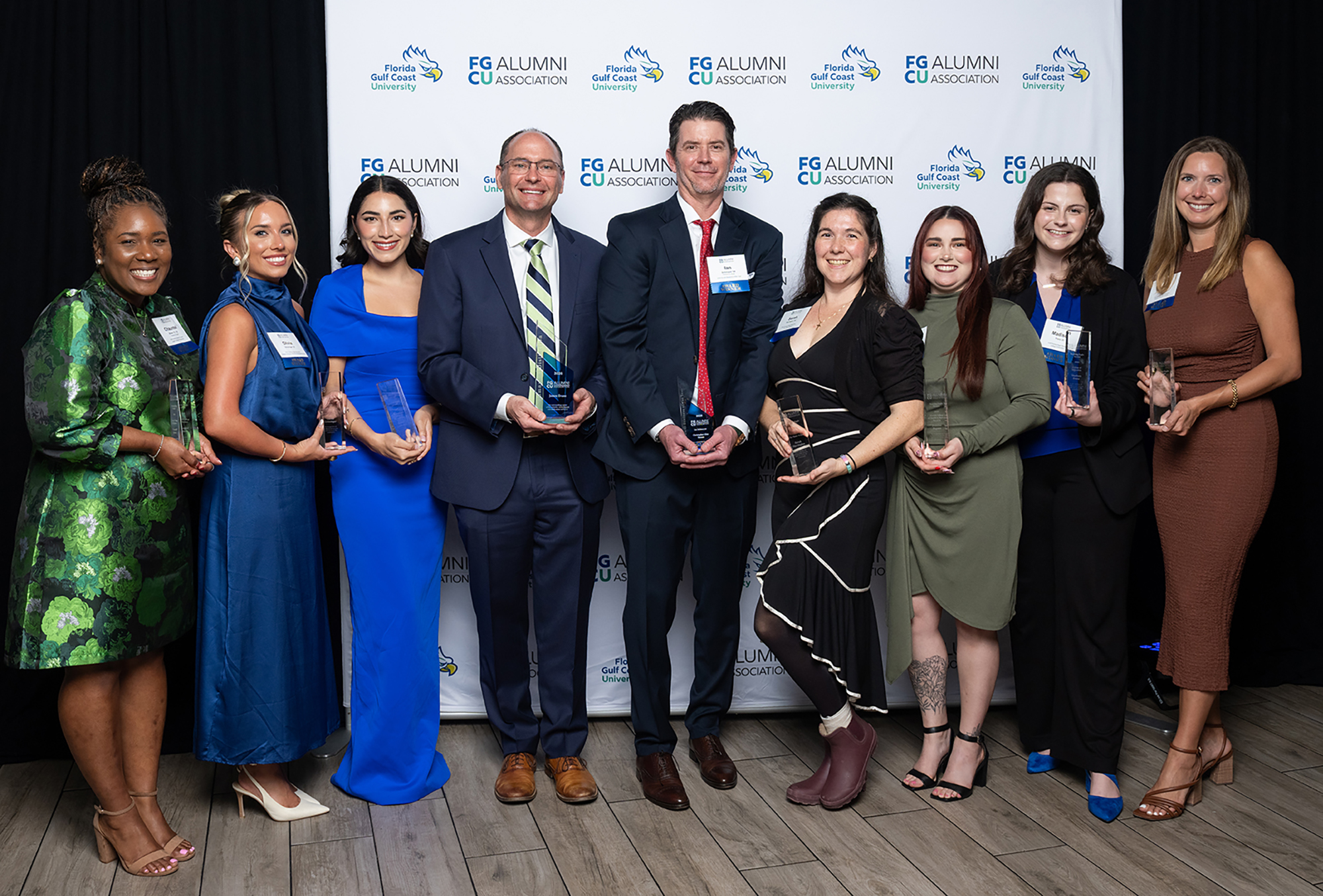 Group of FGCU Alumni Award recipients standing together holding glass awards in front of event backdrop.