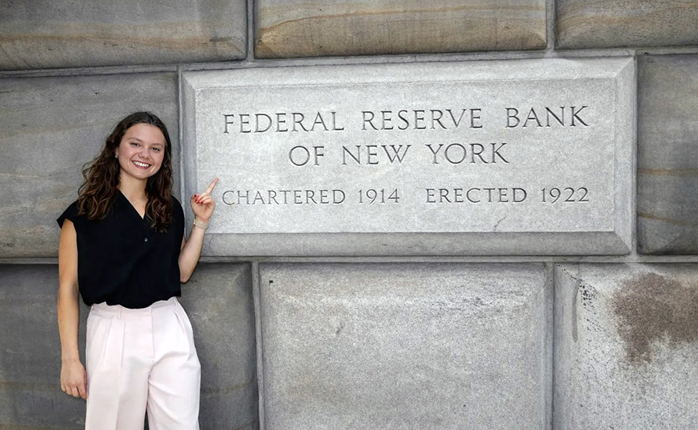Person standing beside the Federal Reserve Bank of New York sign on a stone wall.