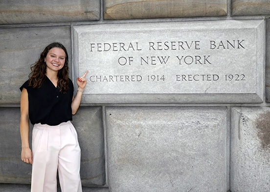 Person standing beside the Federal Reserve Bank of New York sign on a stone wall.