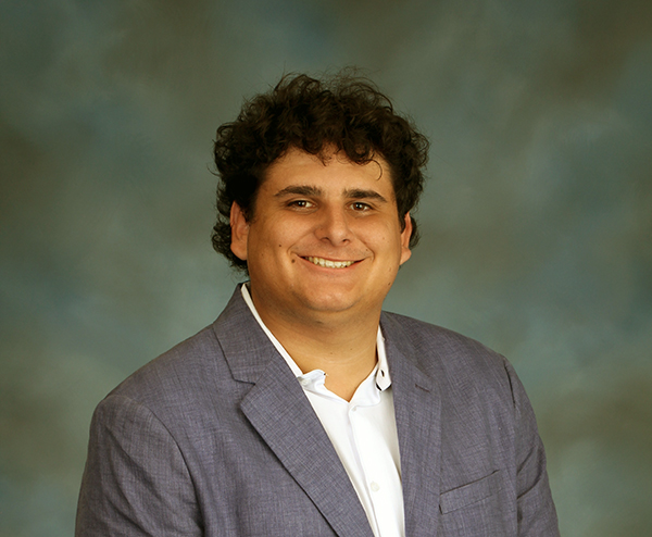 Person wearing a gray blazer and white shirt posed against a studio backdrop.