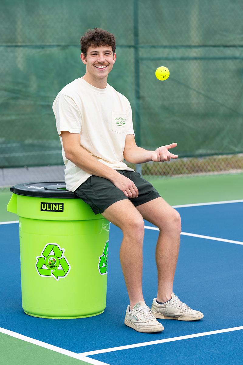 Person sitting on a green pickleball recycling bin while tossing a yellow pickleball.