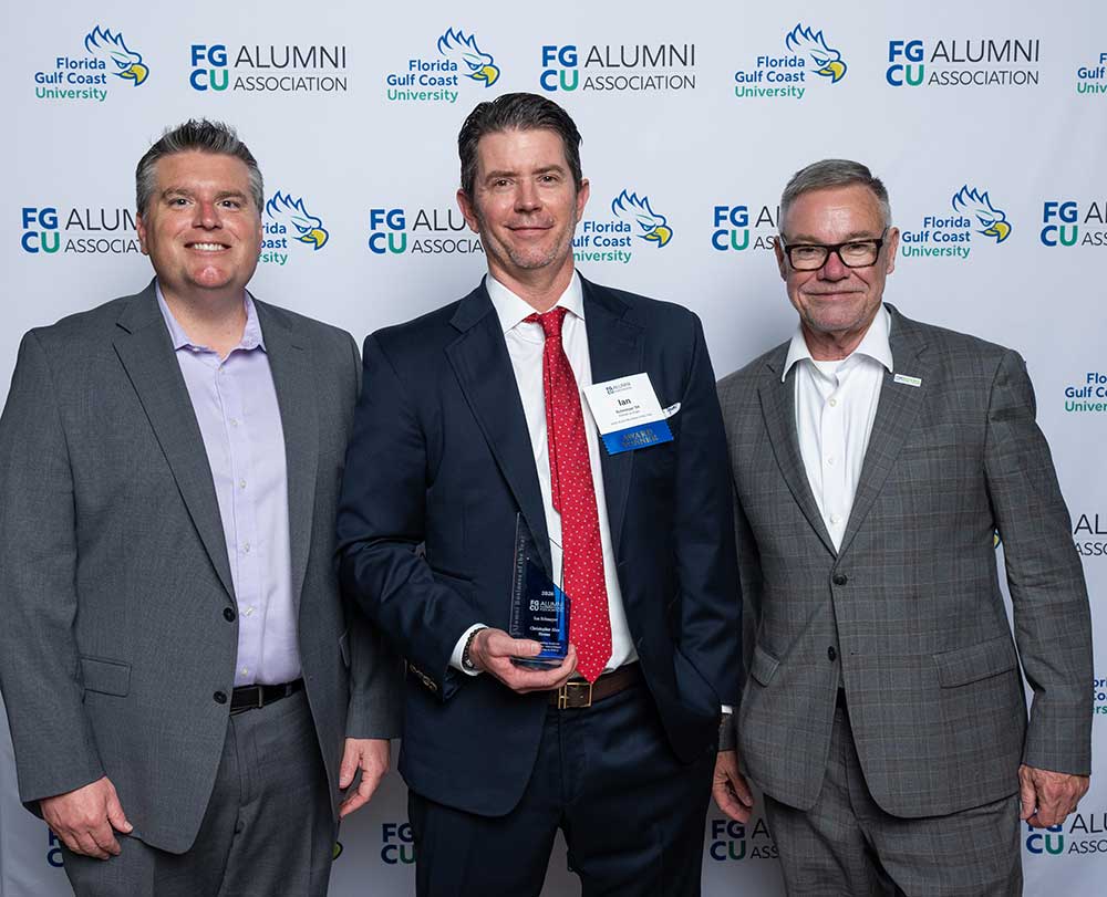 Three people in business attire stand together, one holding an award, in front of an FGCU Alumni backdrop.