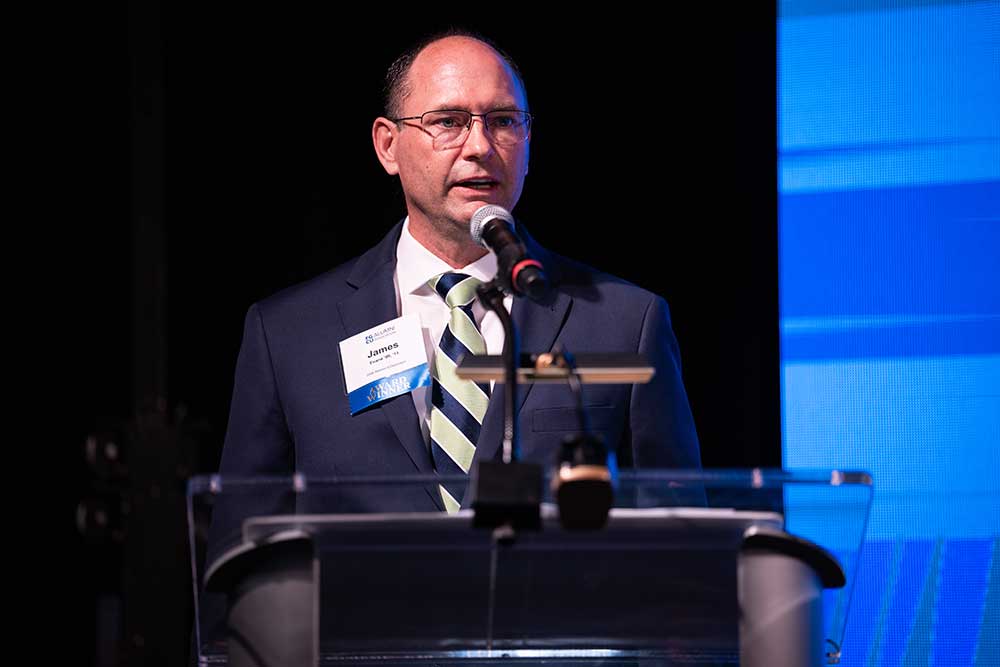 Speaker at a podium addresses the audience beside a large blue event screen.