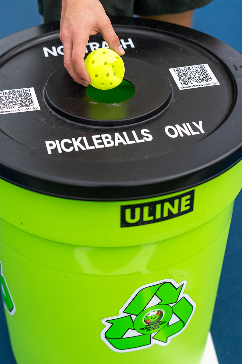 Hand placing a yellow pickleball into a green container labeled Pickleballs Only.