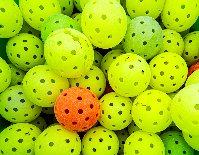 Pile of bright pickleballs, mostly yellow with one orange ball in the center