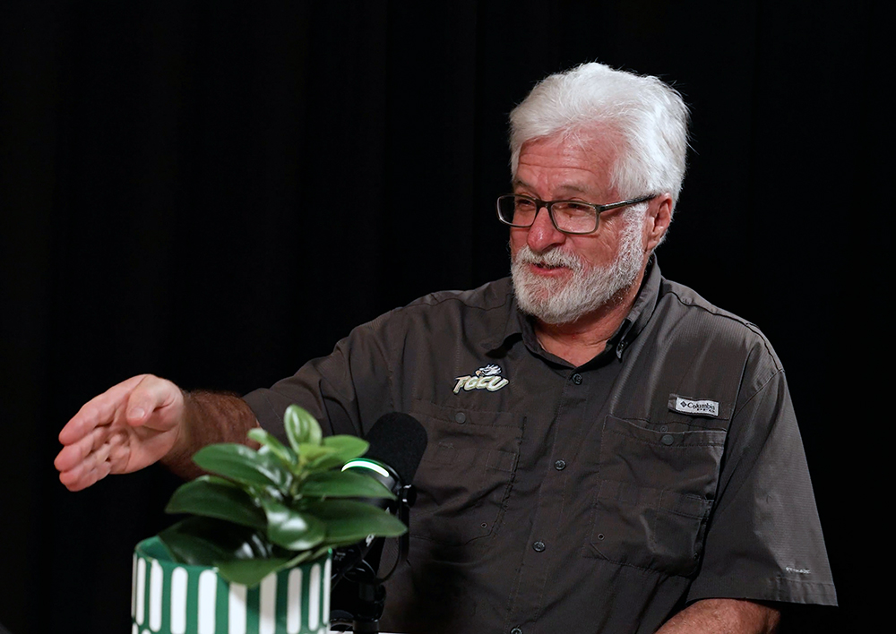 Person speaking into a podcast microphone with a small plant in the foreground.