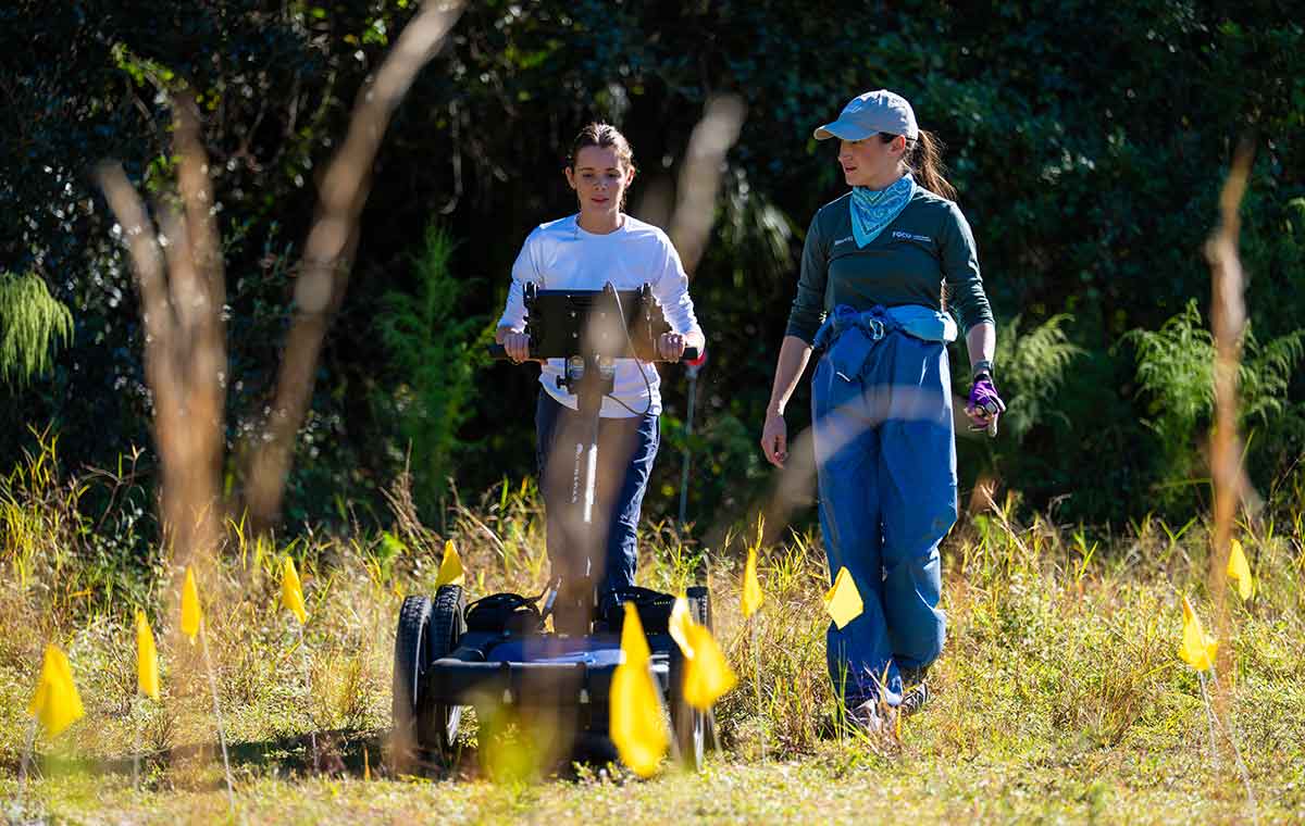 Two women walking in a field. The woman on the left pushes a ground penetrating radar unit that looks like a lawnmower on the bottom with a small screen or monitor at chest-level