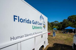 A white truck with the logo for Florida Gulf Coast University and the words Human Identity and Trauma Analysis printed on the side is parked outdoors with blue tents and people walking around