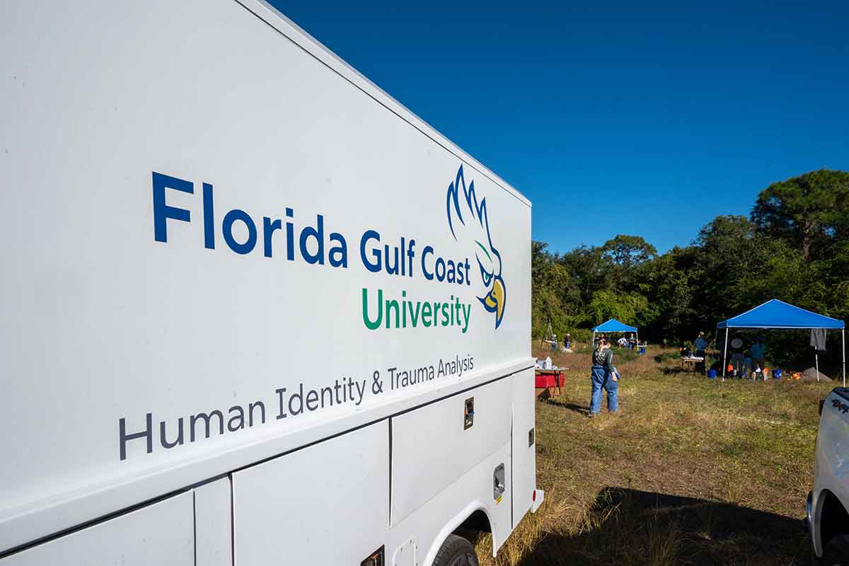 A white truck with the logo for Florida Gulf Coast University and the words Human Identity and Trauma Analysis printed on the side is parked outdoors with blue tents and people walking around