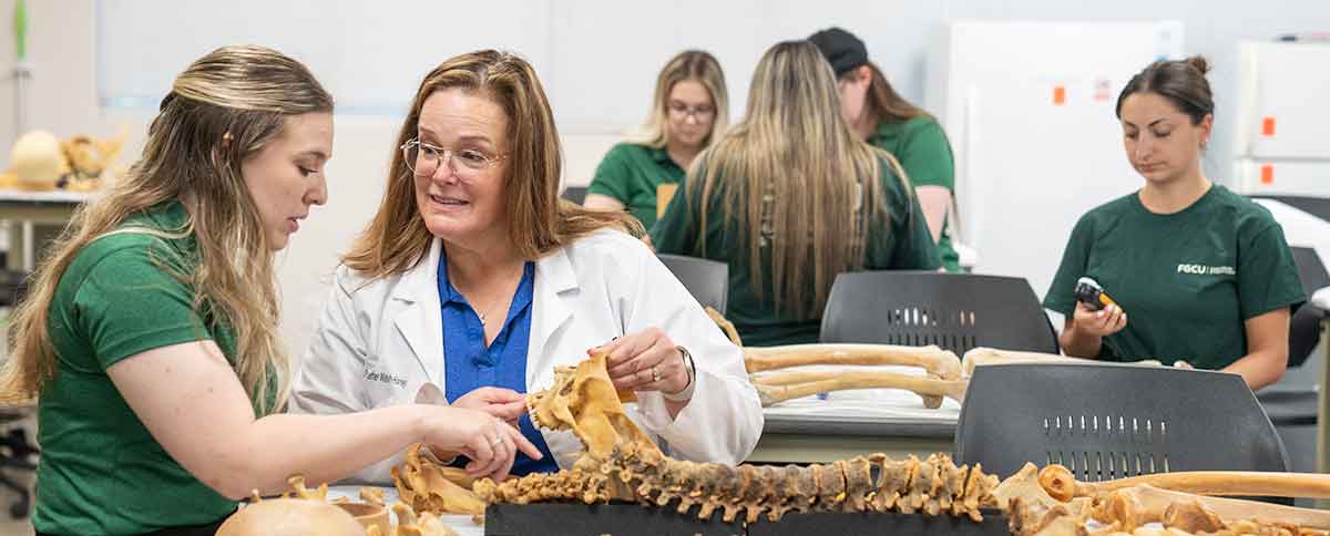Students in a classroom with a woman in a white lab coat, blue shirt and glasses, with a human skeleton laid out before them