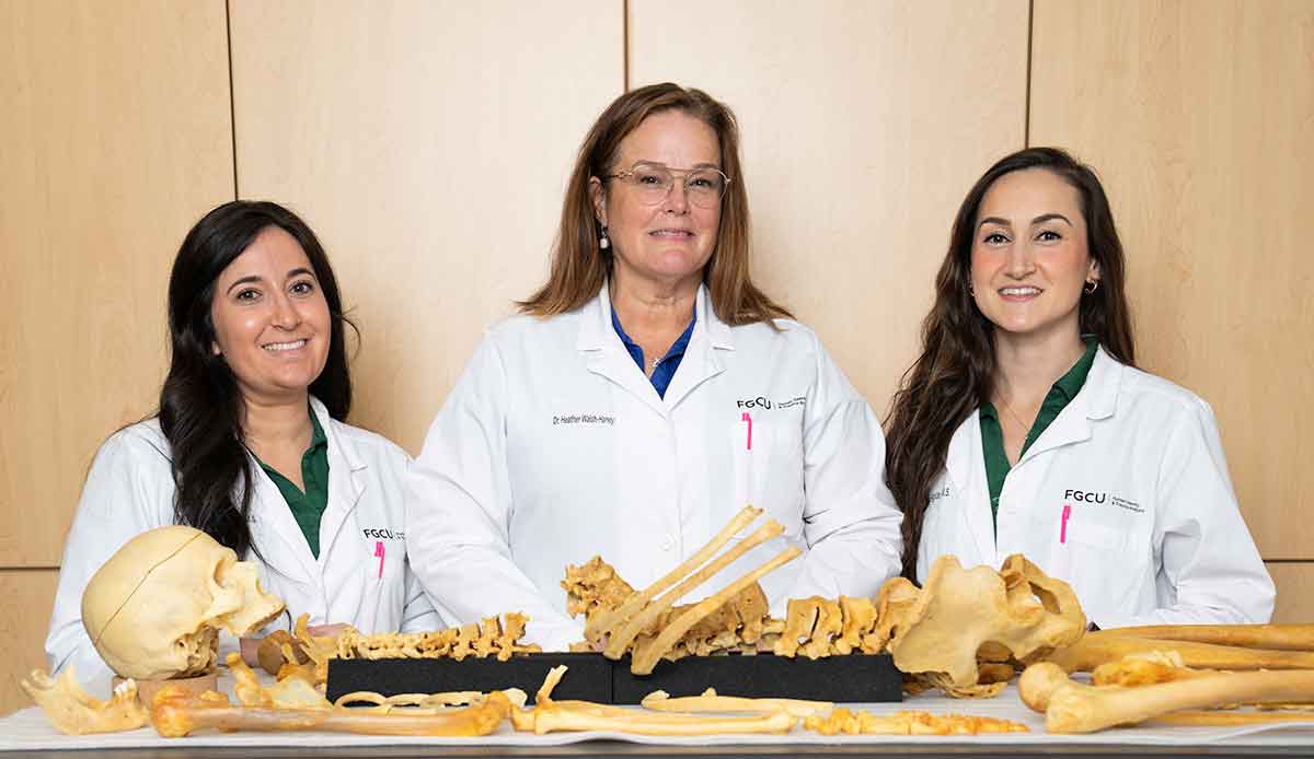 Three women in white lab coats stand in front of a human skeleton