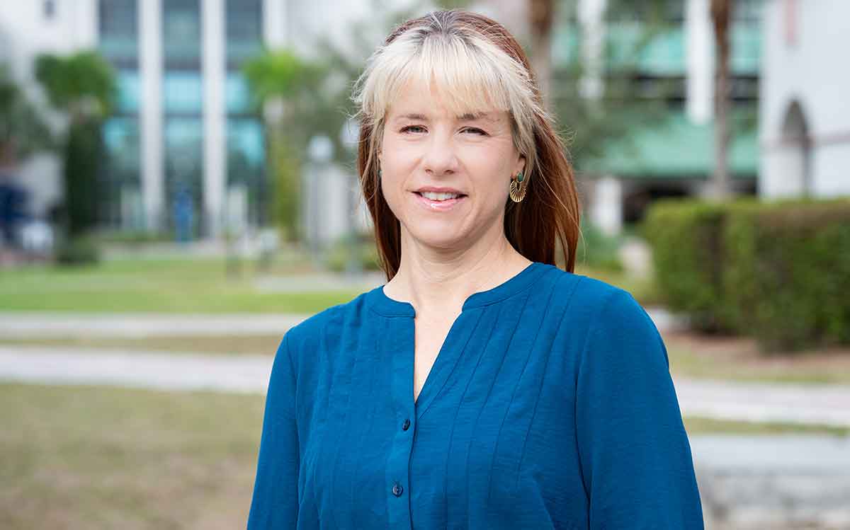 A woman with pale blonde bangs and red hair wearing a blue blouse and smiling outdoors