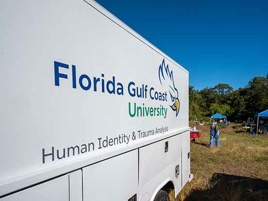 A white truck with the logo for Florida Gulf Coast University and the words Human Identity and Trauma Analysis printed on the side is parked outdoors with blue tents and people walking around